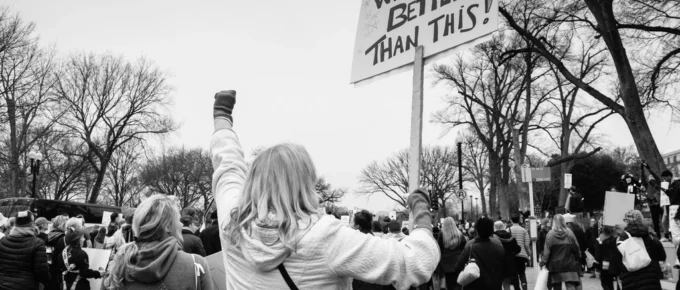 Woman with raised fist at a women's march beside a sign reading We Are Better Than This